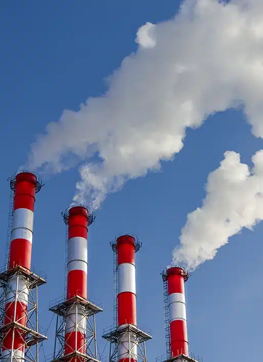 Four red and white smokestacks emit thick clouds of white smoke against a clear blue sky.