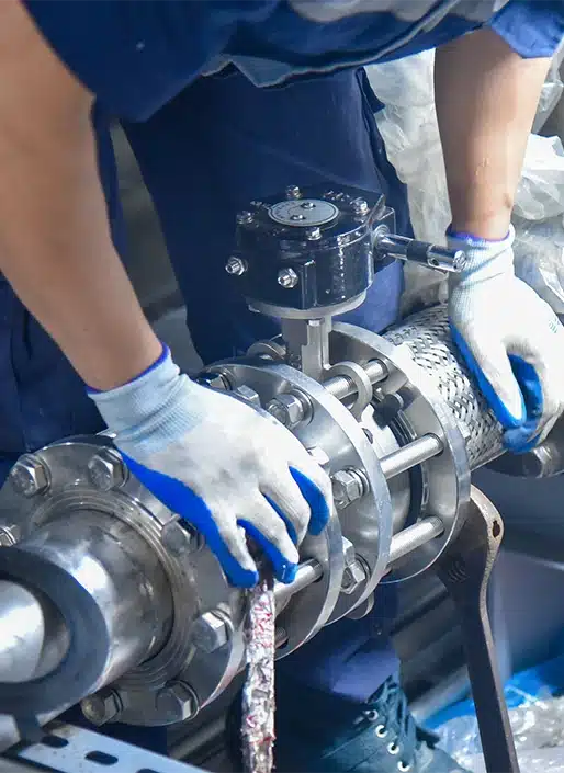 Un ouvrier portant des gants règle une vanne ou un tuyau métallique industriel. Le décor suggère un environnement technique ou mécanique.
