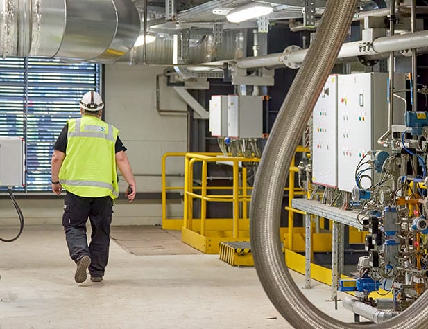 A man in a bright yellow vest and white hard hard is walking away from pipes and control box