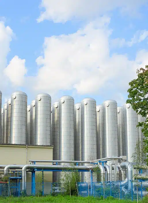 Row of tall, cylindrical metal silos under a blue sky with fluffy clouds. Pipes run along the base, and there is a tree to the right.