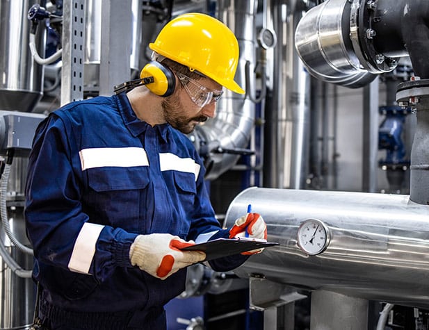 Industrial worker in blue coveralls with a yellow hard hat and earmuffs inspects gauges on machinery, holding a clipboard, in a factory setting.