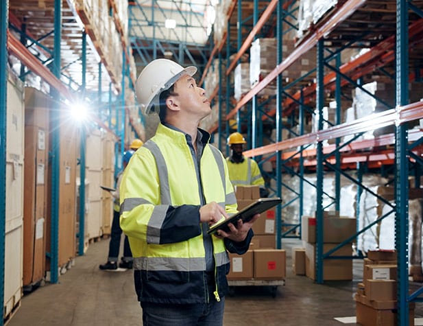 Man in white hard hat and safety reflective jacket, with tabet in hand and looking at large shelves.