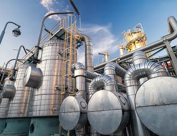 Des tuyaux et des structures métalliques brillants s'entrelacent dans une usine industrielle, sous un ciel bleu.