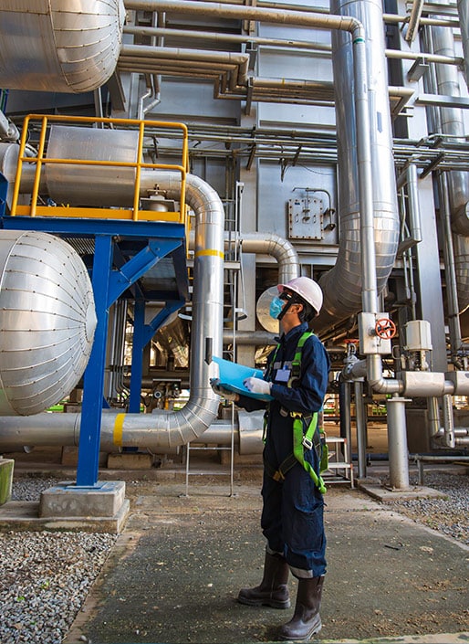 A man in hard hat has a clipboard in his hand and is looking up at tanks and pipes