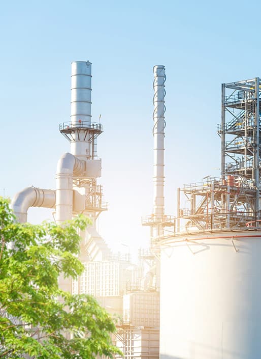 Industrial plant with large metal towers and pipes against a clear blue sky, partially obscured by green trees.