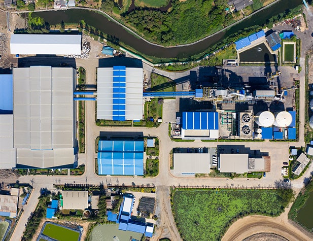 Aerial view of an industrial complex with large blue-roofed buildings, roads, and greenery.