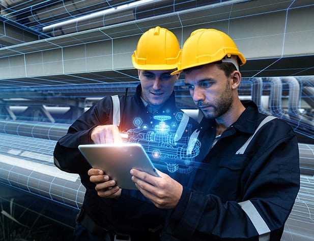 Two engineers in yellow hard hats examine a digital hologram on a tablet. They're in an industrial setting