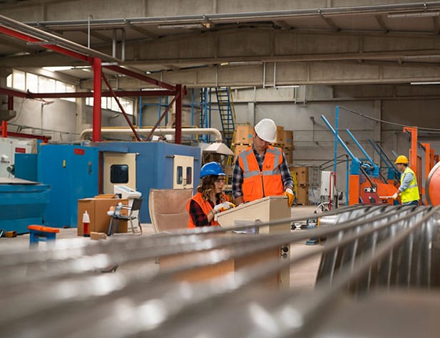Industrial setting with three workers in hard hats and vests. Two review equipment controls, while one inspects machinery.