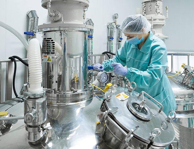 A technician in protective clothing inspects gauges on large, shiny stainless-steel tanks in a sterile industrial setting.