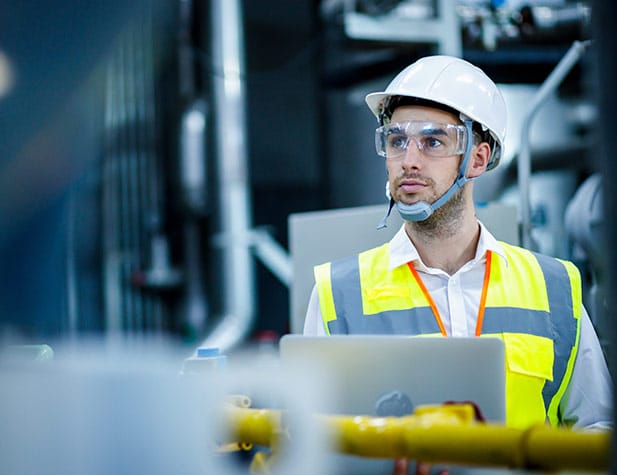 Un homme portant un casque de chantier blanc, des lunettes de sécurité et un gilet jaune réfléchissant, le regard perdu au loin.