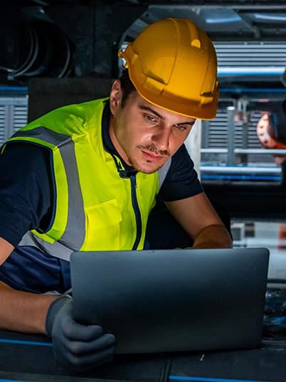 A worker with a yellow hard hat, reflective vest and gloves looking at a computer.