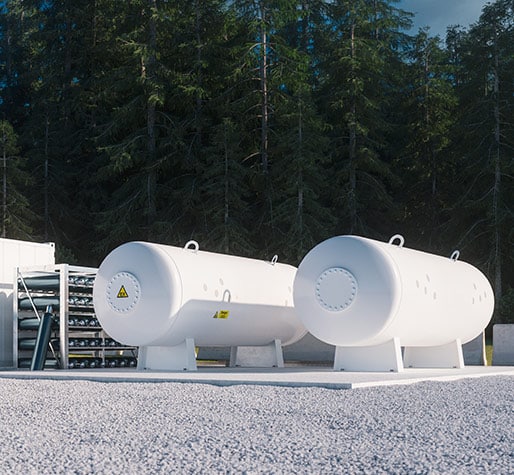 Two large white cylindrical storage tanks on a gravel surface, set against a backdrop of dense evergreen trees.