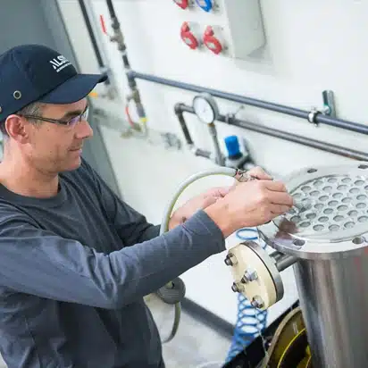 Dans un atelier, un ouvrier portant une casquette noire et des lunettes examine avec une grande attention une machine industrielle comportant un élément métallique circulaire.